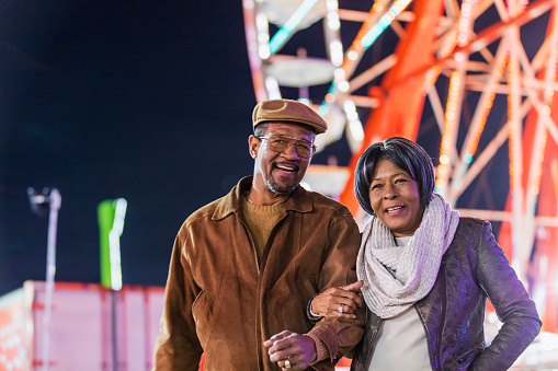 A senior African-American couple in their 60s having fun together at a traveling carnival. An amusement park ride is out of focus in the background.