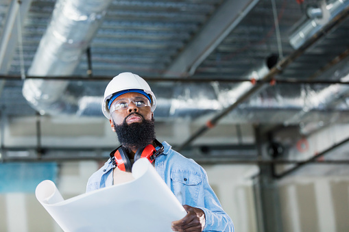 A construction worker wearing a hard hat and safety glasses at an indoor construction site, looking at floor plans. He is a mature African-American man in his 40s with a thick beard. The structure being built is an office building.