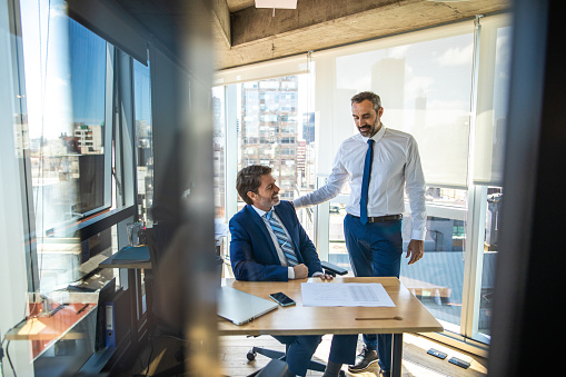 Two successful, formal dressed businessmen having a pleasant conversation in brightly lit modern office