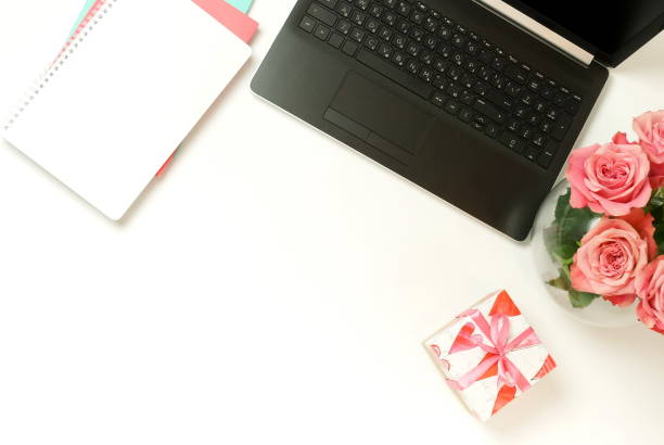 istock top view women's office desk on  white background selective focus. Female workspace with laptop, beautiful pink roses flowers in a vase, gift box, accessories, notebook mock up, glasses. 1207310415