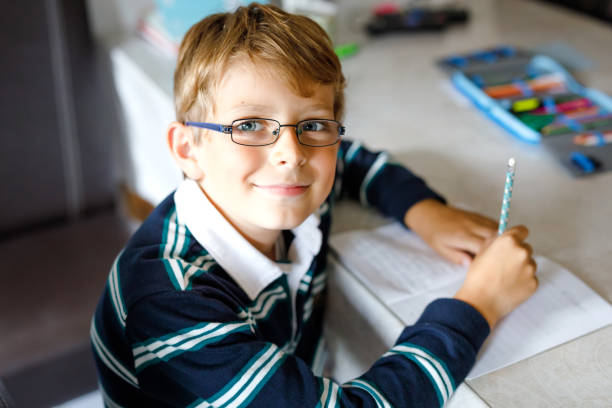 Thoughtful little kid boy with glasses at home making homework, writing text with colorful pens. Little child doing exercise, indoors. Elementary school and education.