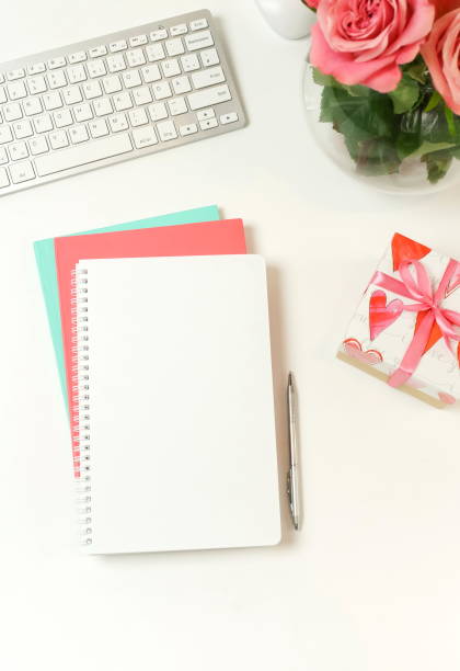 istock top view women's office desk on  white background selective focus. Female workspace with laptop, beautiful pink roses flowers in a vase, gift box, accessories, notebook mock up, glasses. 1207311762