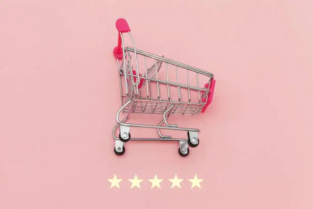 Photo of Small supermarket grocery push cart for shopping toy with wheels and 5 stars rating isolated on pastel pink background. Retail consumer buying online assessment and review concept