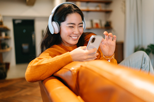 A stylish young woman is sitting on a sofa and listening to music with a wireless bluetooth headphones connected to her smart phone in the living room at home.