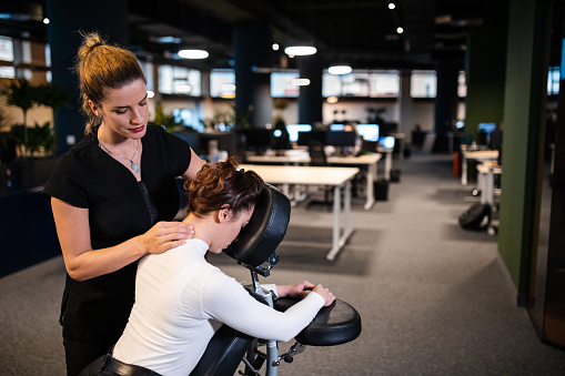 Female massage therapist giving businesswoman massage for her back and neck pain, while she sitting in a mobile massage chair in her work  office