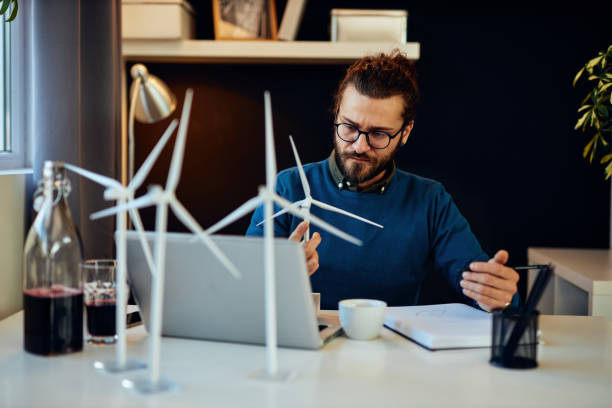 istock Young creative caucasian bearded environmentalist  with curly hair sitting in his office, holding windmill model and drawing new better model in notebook. 1207343558