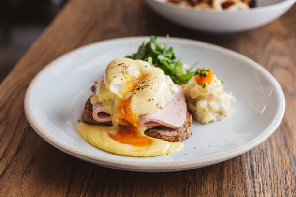 istock Close-up Eggs Benedict with ham that cut and show filmy yolk and cheese, toast and mashed potato. Served with salad on a white plate on wooden table. 1207343967