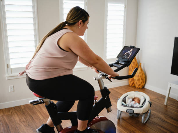 young mother using exercise bike in a home - mother exercising baby dieting imagens e fotografias de stock