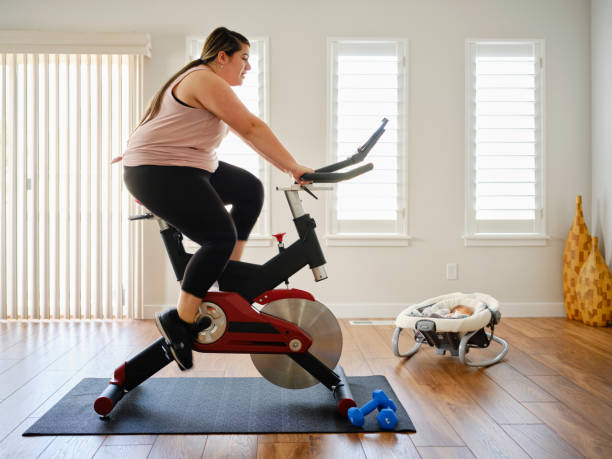 young mother using exercise bike in a home - mother exercising baby dieting imagens e fotografias de stock
