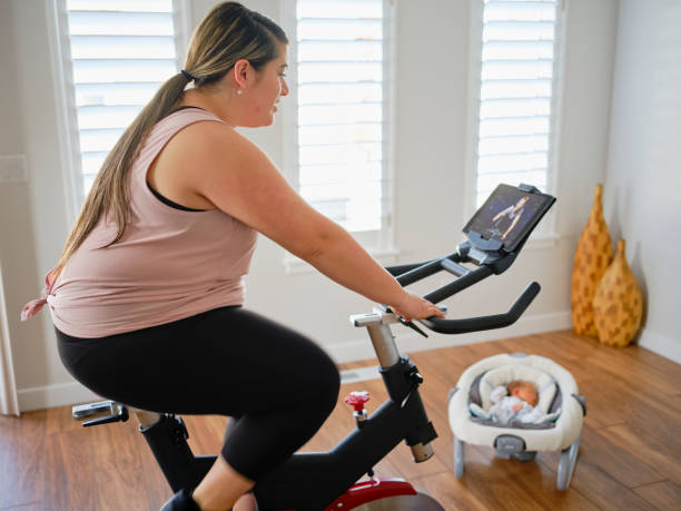 young mother using exercise bike in a home - mother exercising baby dieting imagens e fotografias de stock