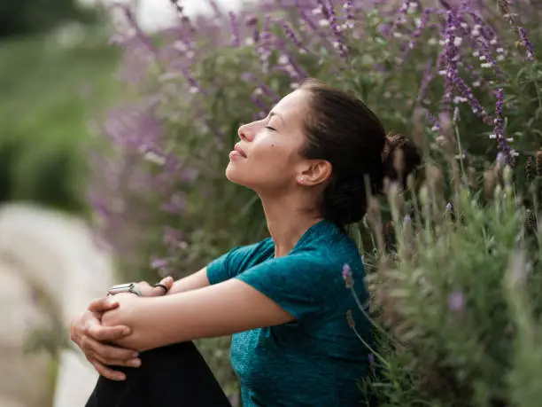 A latin woman sitting with her eyes closed and relaxing next to some flowers.