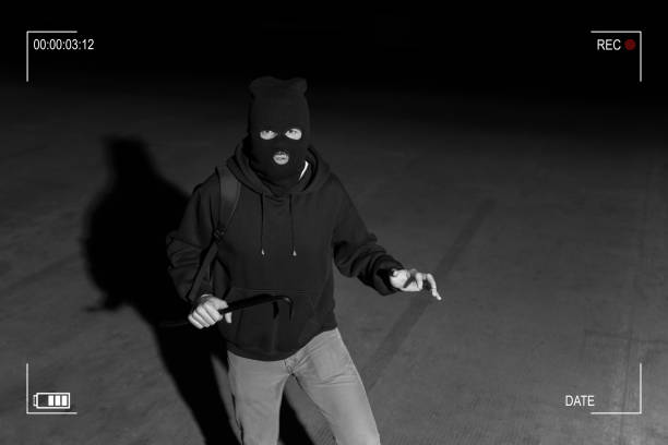 CCTV View Of Thief Standing In Dark Alley Surveillance camera caught burglar in ski mask holding crowbar while making eye contact in dark parking lot burglary stock pictures, royalty-free photos & images
