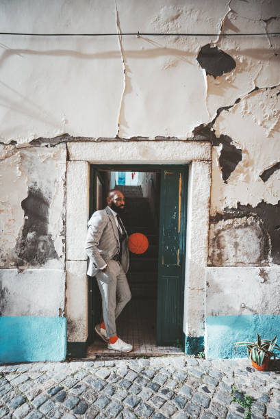 A stylish fancy African man with a beard is standing in the doorway with a basketball in hands; black elegant guy win a plaid costume in the doorway with a flacked wall outside and dark corridor abaft
