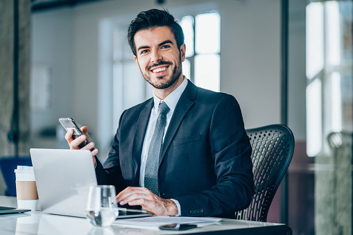Shot of a young businessman sitting on desk in modern office and looking at camera. Portrait of a handsome businessman working on laptop and using smart phone in the office.