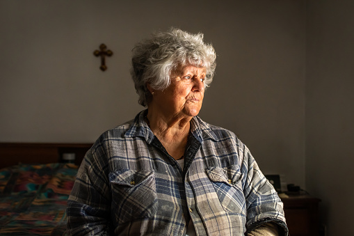 Portrait of an elderly Portuguese woman sitting on a bed by a window in her room.