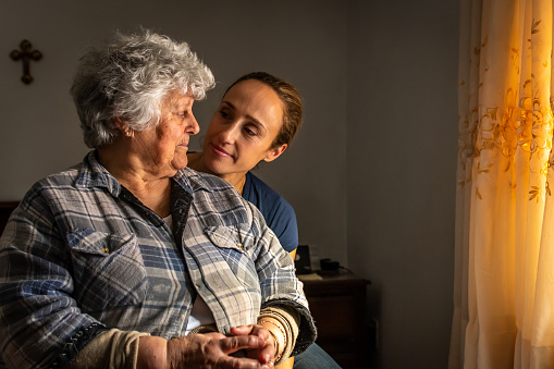 Portrait of a daughter holding her elderly mother, sitting on a bed by a window in her mother's room.