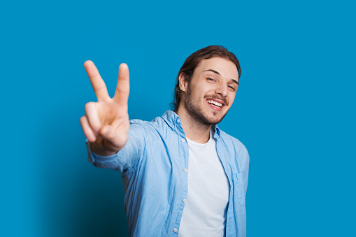 Smiling unshaven caucasian man with long hair dressed in a t-shirt and a blue shirt is gesturing the victory sign a blue background
