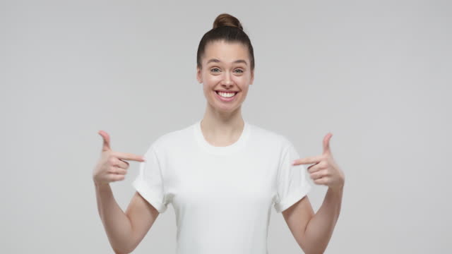 Young girl pointing to her white t-shirt with thumbs up gesture, showing empty space for your text or logo, isolated on gray background