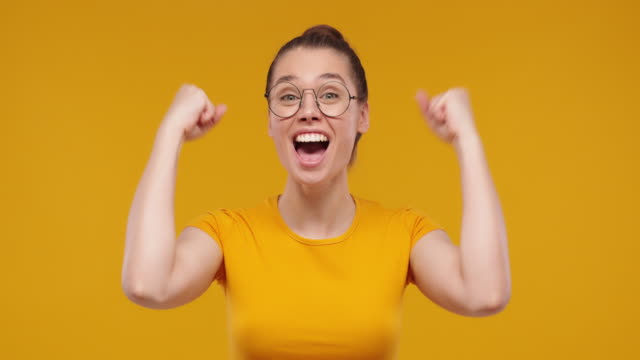 Young pretty fan girl shouting while her team win, raised both fists in victory gesture, isolated on studio yellow background