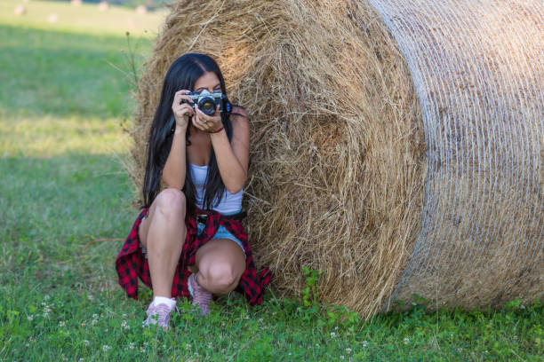 Yоung woman with vinatge camera on the field taking photos near a pile of hay in sunny summer day