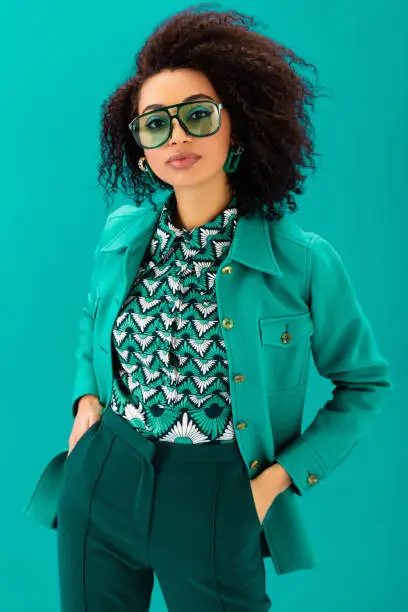 Photo of african american woman in jacket with hands in pockets looking at camera isolated on turquoise