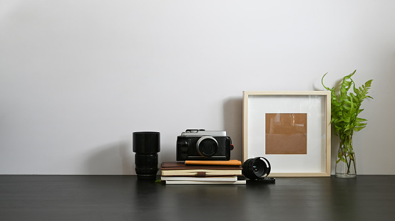 Photographer workspace with white wall as background. Camera, Lens, Stack of books, Picture frame and Potted plant putting together on black wooden table in orderly living room.