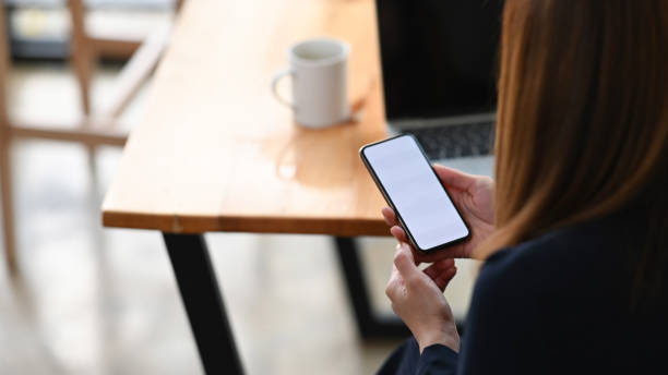 istock Cropped image of charming woman working as project manager using/holding cropped black smartphone with white blank screen in hands while seated at the wooden working table. 1207479298