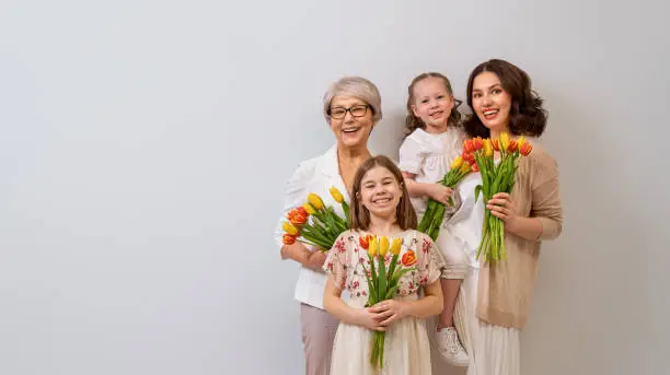 Happy women's day! Children daughters are congratulating mom and grandma giving them flowers tulips.Granny, mum and girls smiling on light grey background. Family holiday and togetherness.