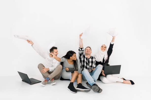 istock Multiethnical university students, business partners, sitting together at the floor, using laptops, working on new startup, happy and satisfied and keeping arms raised 1207487336