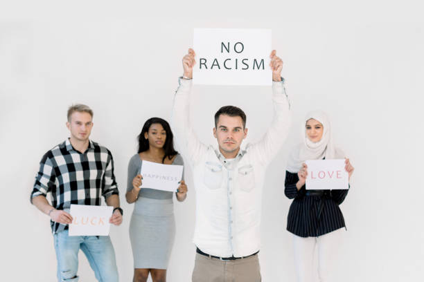 istock Handsome Caucasian young man protests with a poster, No racism concept, together with three multiethnic friends, African and Muslim women and Caucasian man. Isolated on white 1207487405