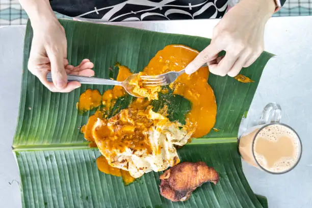Photo of Person eating roti canai or paratha with curry on banana leaf with teh tarik or milk tea.