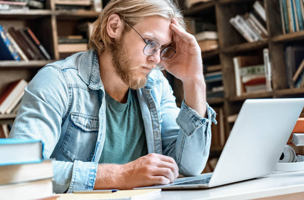 istock Young concentrated man student home library desk read look at modern laptop pc screen hold hand at head skype course web study online training e learning take test distant electronic education concept 1207490402