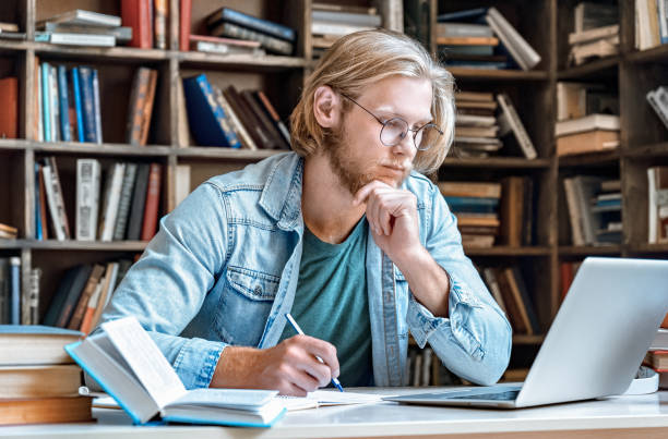 istock Focused young man student hipster home library desk read look at modern laptop pc screen make notes has skype course web study online training e learning take test distant electronic education concept 1207490418