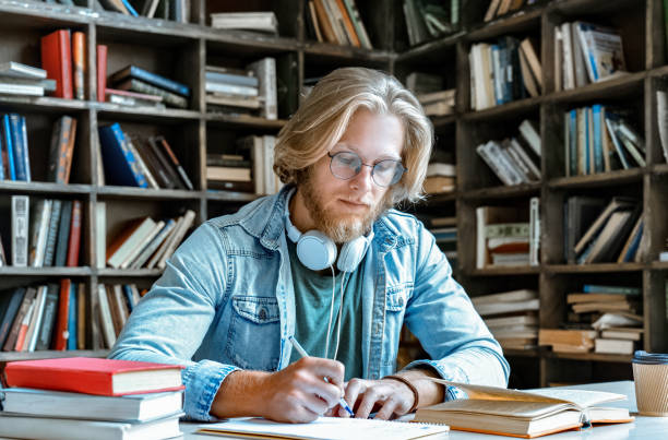 istock Concentrated bearded young man student teacher hipster glasses sit study library desk hold pen make notes from book hand write essay notebook fill in journal do homework make report education concept 1207490432