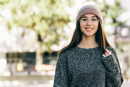 Portrait of young woman smiling, wearing transparent eyeglasses, gray sweater and pink hat. Outdoor image of pretty student girl looking at the camera and relaxing in the city street.