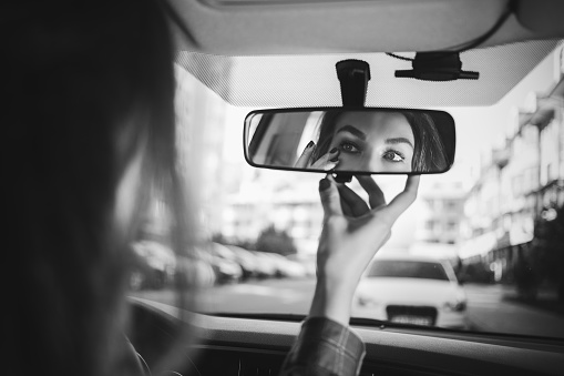 Young woman looking in rear view mirror and putting make up in car. modern busy life.beautiful girl sitting in drivers seat of car doing make up, applying lipstick.