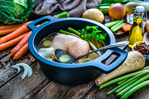 High angle view of a cooking pan filled with raw chicken bouillon ingredients shot on rustic kitchen table. Ingredients for preparing chicken broth like raw chicken, carrots, kale, potatoes, celery and onions are also all around the pan. High resolution 42Mp studio digital capture taken with SONY A7rII and Zeiss Batis 40mm F2.0 CF lens
