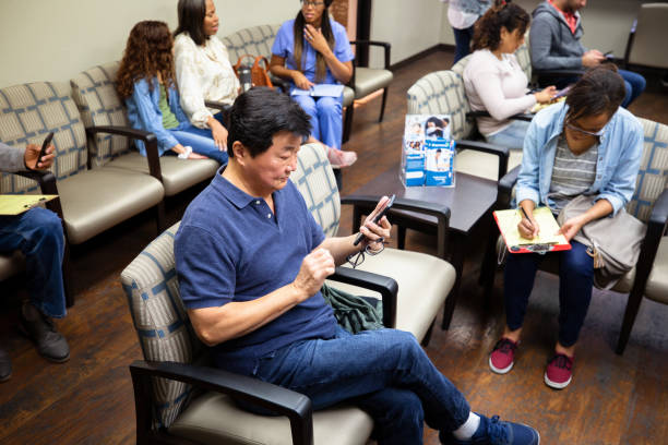 Asian senior man waits for doctor appointment An Asian senior man uses his smartphone to pass the time while waiting in a medical clinic's waiting room. waiting room stock pictures, royalty-free photos & images