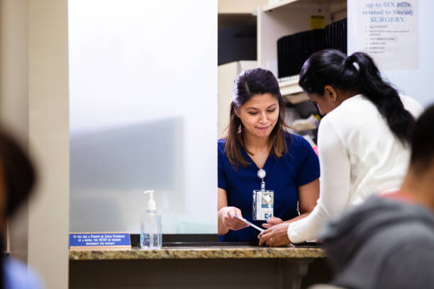 Mid adult woman checking in for medical appointment A mid adult Indian woman talks with a nurse at the window at a medical clinic. They are reviewing new patient paperwork. receptionist stock pictures, royalty-free photos & images