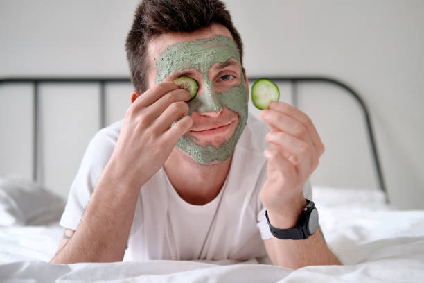 joven con una camisa blanca con máscara cosmética verde aplicada sosteniendo trozos de pepino - facial mask spa treatment cucumber human face fotografías e imágenes de stock