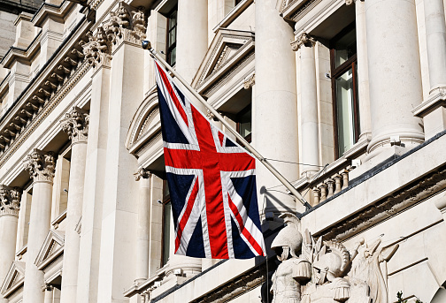 Image shows a view of Union Jack flag on a flagpole in front of building during day in London, England
