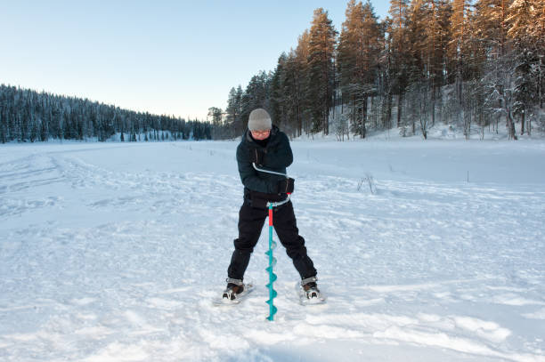 Man hole drilling ice Finland, Lapland, Salla. February 2020. The man is on the polar circle in his vacation. The man is wearing warm clothing. On his snowshoes he is standing on the frozen lake. There is a lot of snow on the ice. With the long ice drill, the drill turns in the ice. This creates a hole in the ice where he can go fishing. man ice fishing stock pictures, royalty-free photos & images