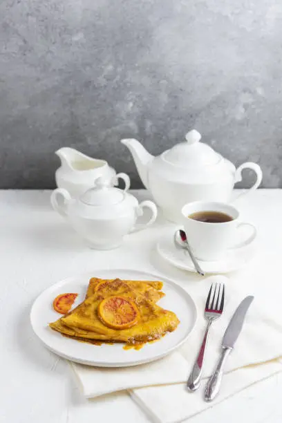 Breakfast, two crepe suzette pancakes in orange sauce and a cup of tea, teapot, sugar bowl, creamer. Vertical still life food on a white table and a concrete wall for a copy space.