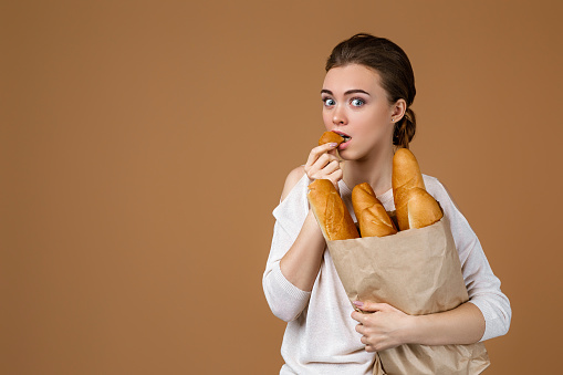 Portrait of beautiful smiling young woman holding paper bag with bread on studio yellow background. girl eating fresh fragrant long loaf. copy space