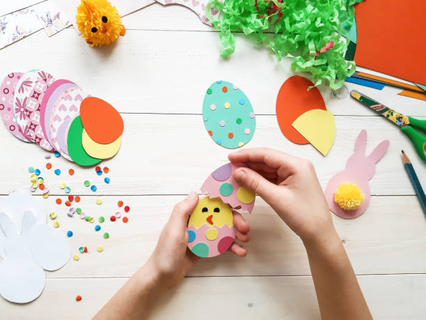 The child makes crafts with his own hands for the Easter. Colorful handmade from multi-colored paper. Scissors, cardboard, eggs, chicken, rabbit. Art creativity on a wooden table. Top view, copy space