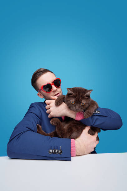 Young man in heart-shaped sunglasses sitting at table and holding displeased cat against blue background