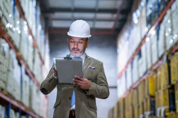 50-year-old man of average age engineer in a free zone company dressed in white safety helmet and formal clothes carries in his hand a table with documents that he is reading very carefully in the back we observe boxes and pallets of the company