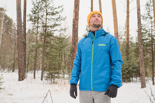 Handsome young man in blue jacket looking around and enjoying winter stroll in forest