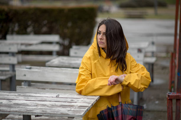 Young woman in yellow raincoat sitting on the bench, holding umbrella and waiting for someone.