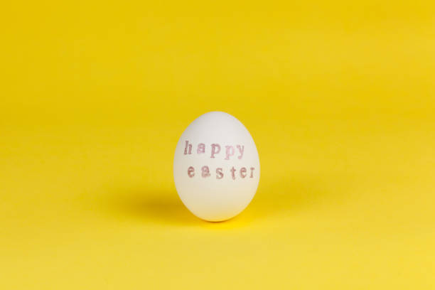 istock One white chicken egg with words Happy Easter written with help of stamps and pink metallic paint on yellow background, copy space. DIY, festive concept. Side view. Horizontal 1207560236
